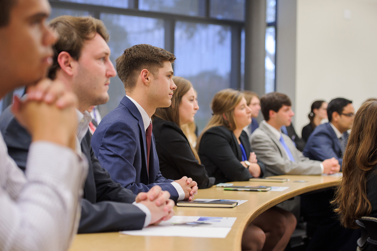 Students in suits sit at a classroom desk. 