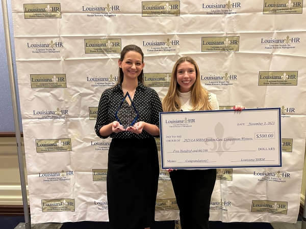 Two women in front of a SHRM banner. One holds a trophy the other holds a large check for $500. 