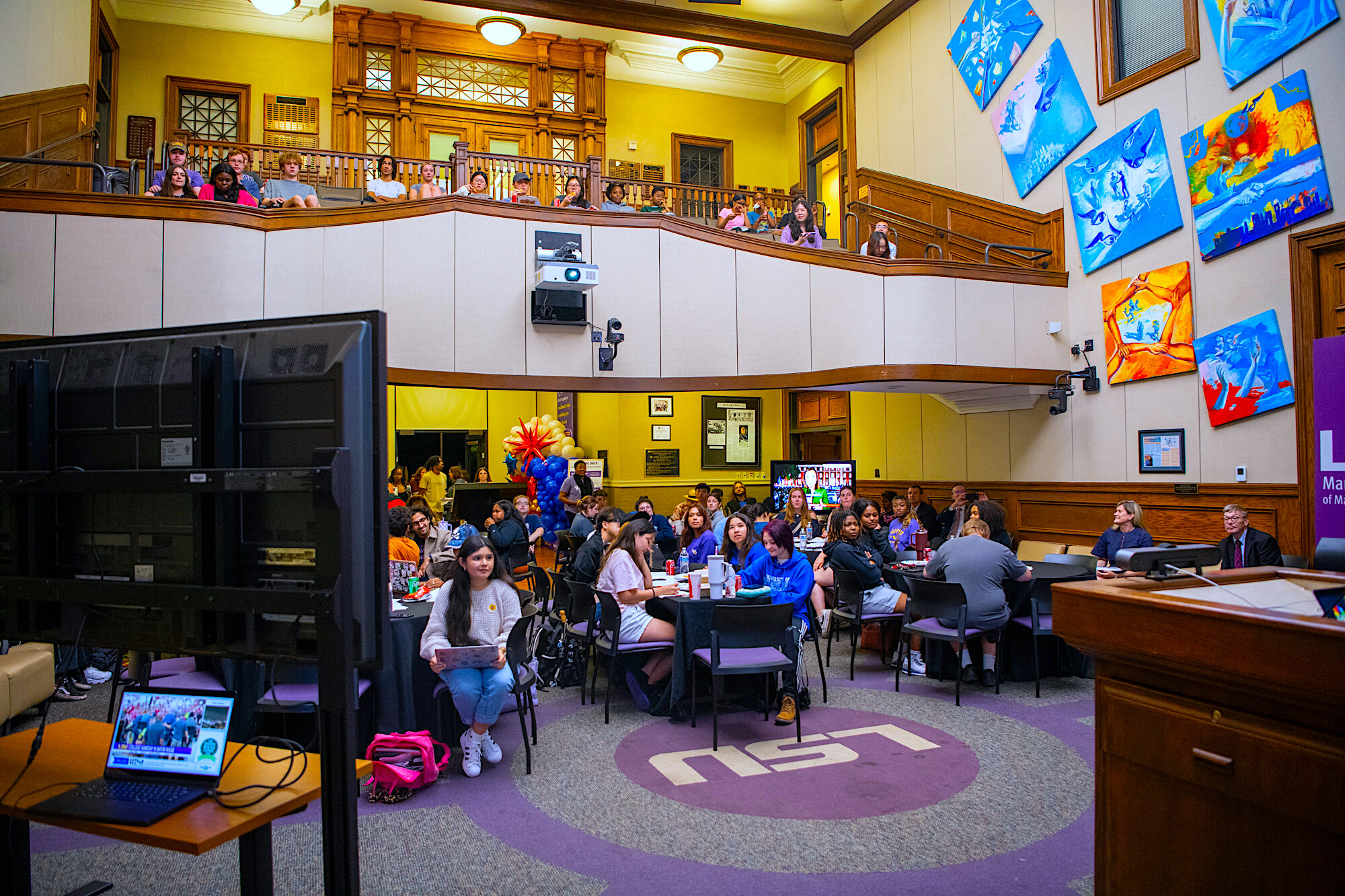 An image of the Holliday Forum showing both the floor and the top viewing row full of students. The floor is filled with tables that the students sit at.