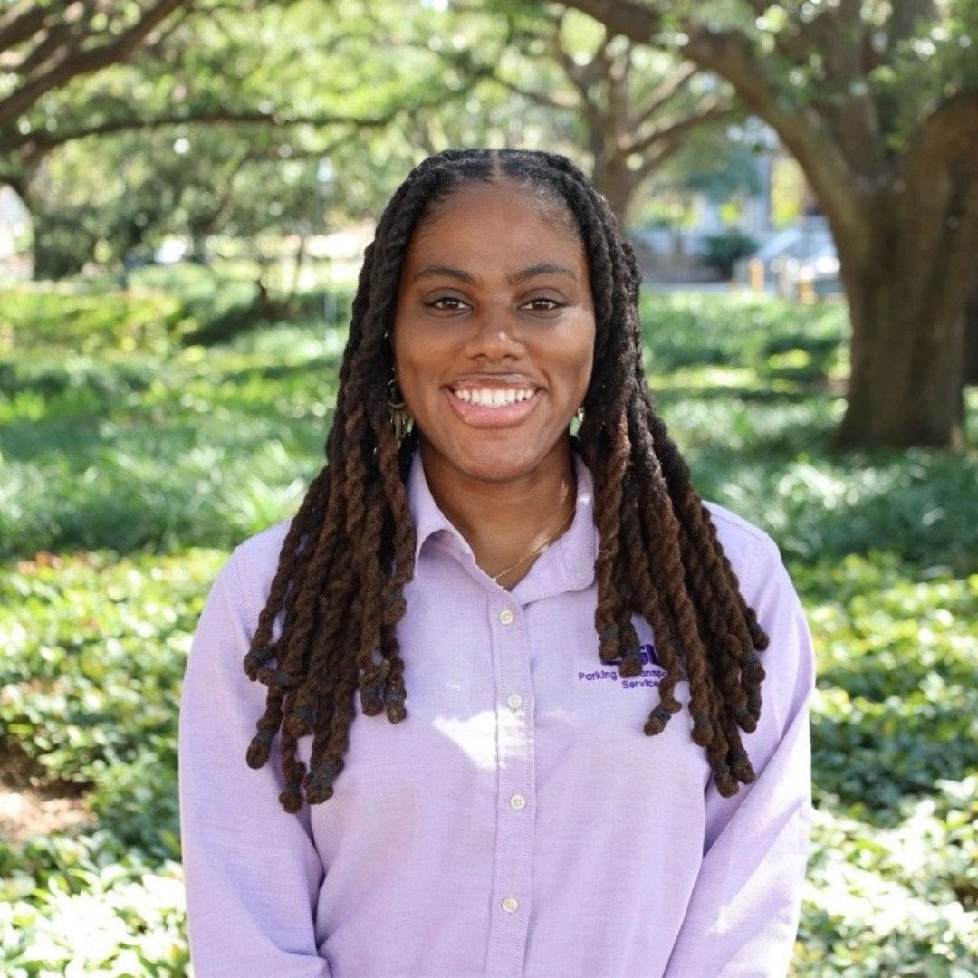 Woman in purple shirt smiling and standing in front of a tree and bushes